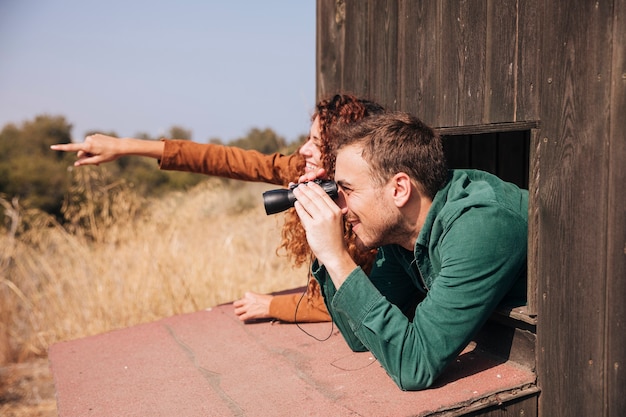 Two birdwatchers looking out over dry grassland from a wooden hut, one with binoculars raised and one pointing