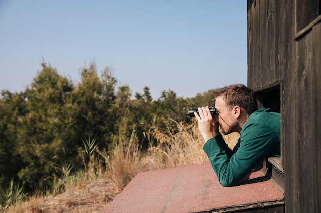 A birdwatcher leaning out of a wooden lookout to scan dry grassland through binoculars
